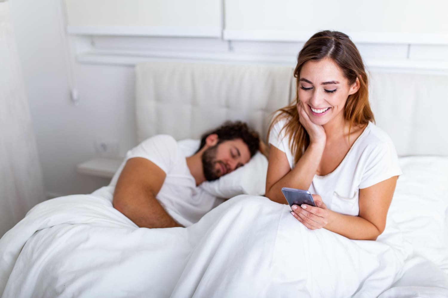 Mulher sorridente usa celular na cama, homem dorme ao lado.