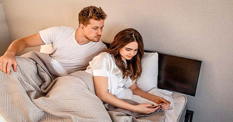 Casal na cama, mulher no celular, homem a observa.