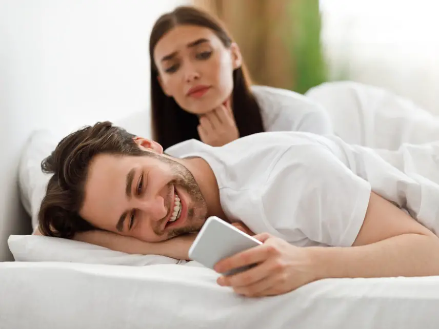 Homem sorridente usa celular na cama, mulher triste observa ao fundo