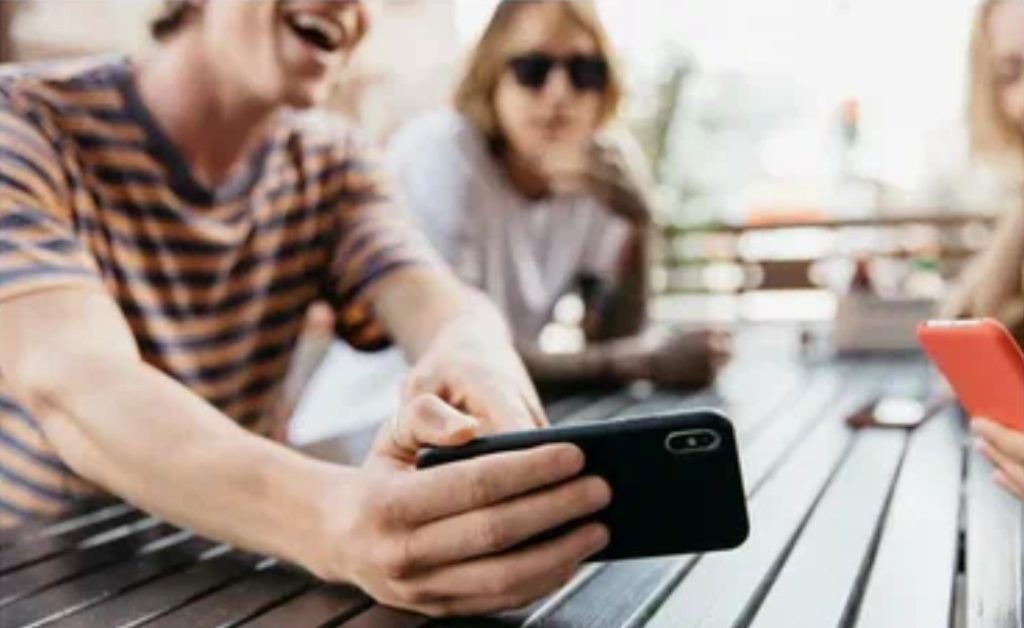 Três amigos usando smartphones ao ar livre, representando a interação social e compartilhamento de conteúdo.
