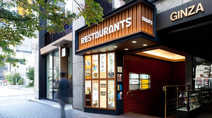 Entrada de um edifício com letreiro \"RESTAURANTS\" e \"SHOPS\", localizado em uma área urbana movimentada.