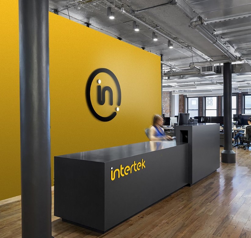 Área de recepção de escritório com parede amarela, logo preto e branco, e balcão preto com letreiro amarelo "intertek".