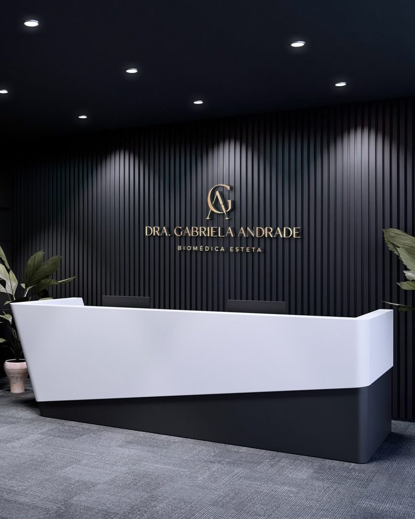 Área de recepção moderna com balcão branco e preto, parede preta com logo \"DRA. GABRIELA ANDRADE BIOMÉDICA ESTETA\" em letras estilizadas.