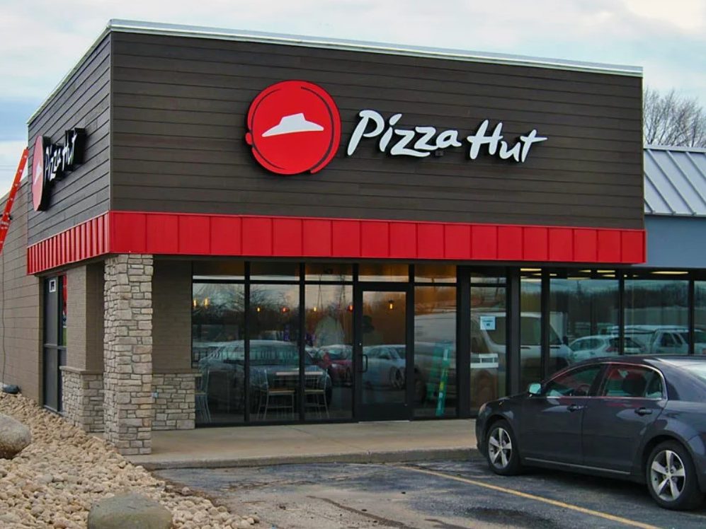 Fachada do restaurante Pizza Hut com logotipo branco em uma fachada vermelha, com design moderno, janelas grandes e reflexivas.