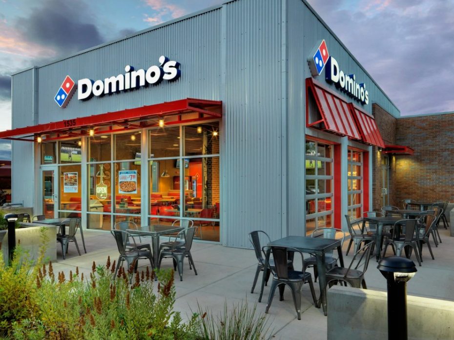 Fachada de um restaurante Domino's Pizza com grandes janelas de vidro e fachada de metal ondulado, com mesas ao ar livre.