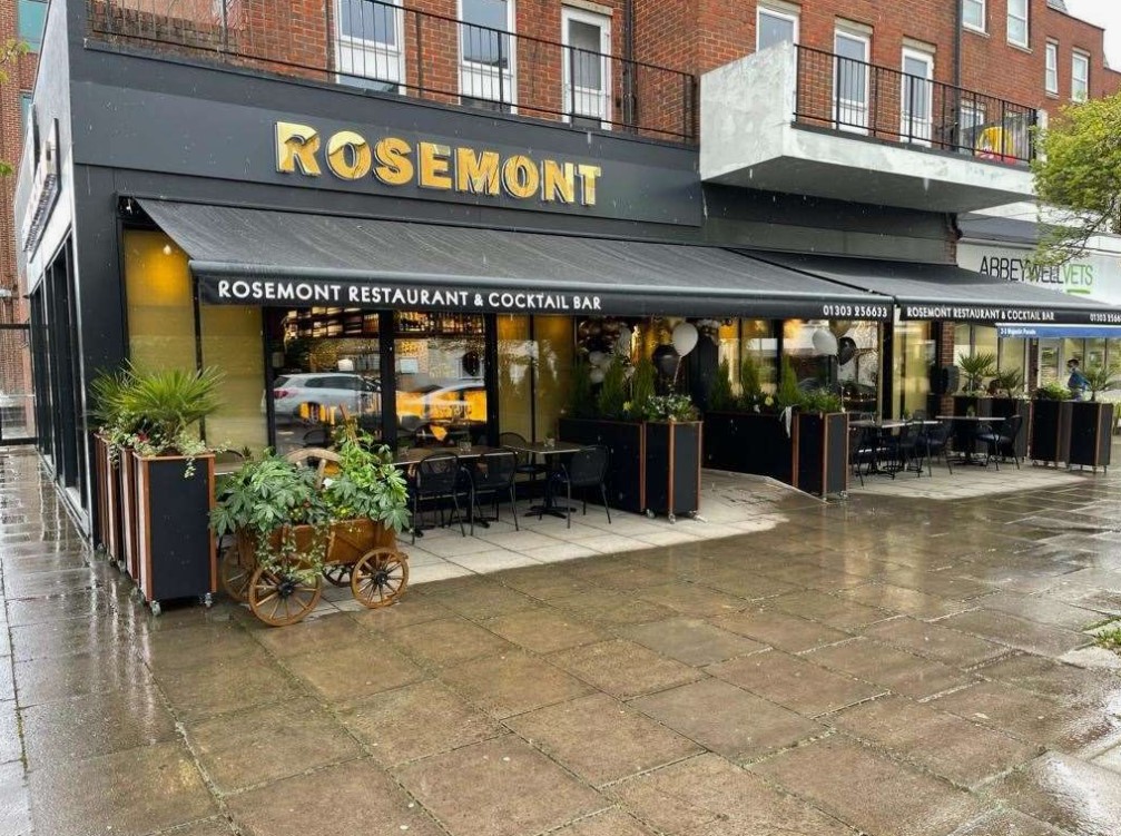 Fachada de um restaurante e bar de coquetéis "Rosemont" com toldo preto, letreiro branco, mesas externas e plantas.