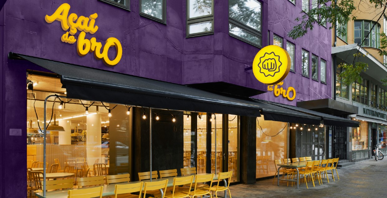 Fachada do restaurante ou café \"Açaí do brO\" com fachada roxa e letreiro amarelo, além de área de estar ao ar livre.