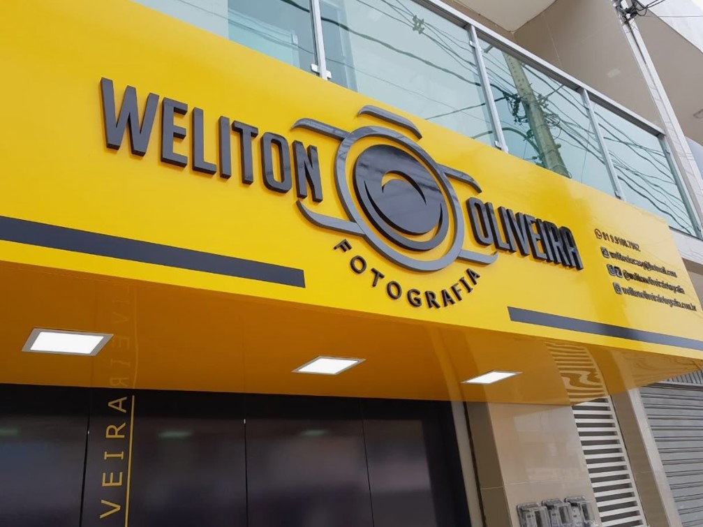 Fachada de uma loja de fotografia com letreiro amarelo e preto, nome \"Weliton Oliveira Fotografia\", e logo de câmera estilizado.