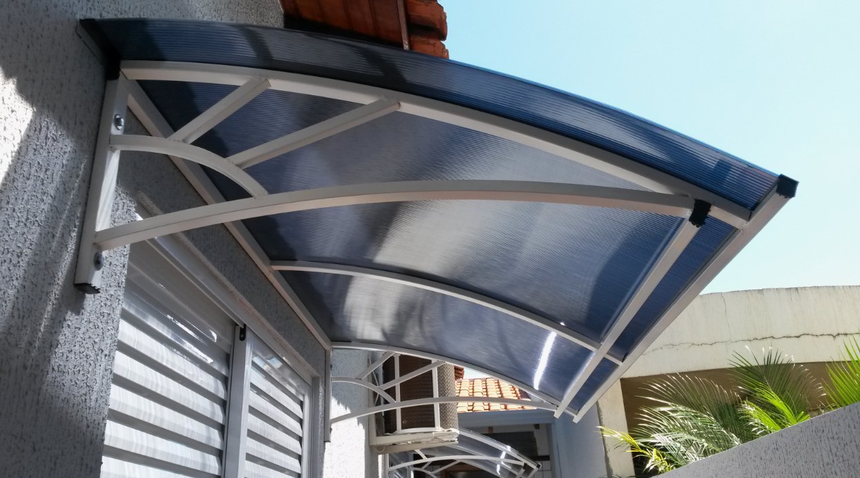 Toldo curvo de policarbonato sobre janela com estrutura metálica branca.