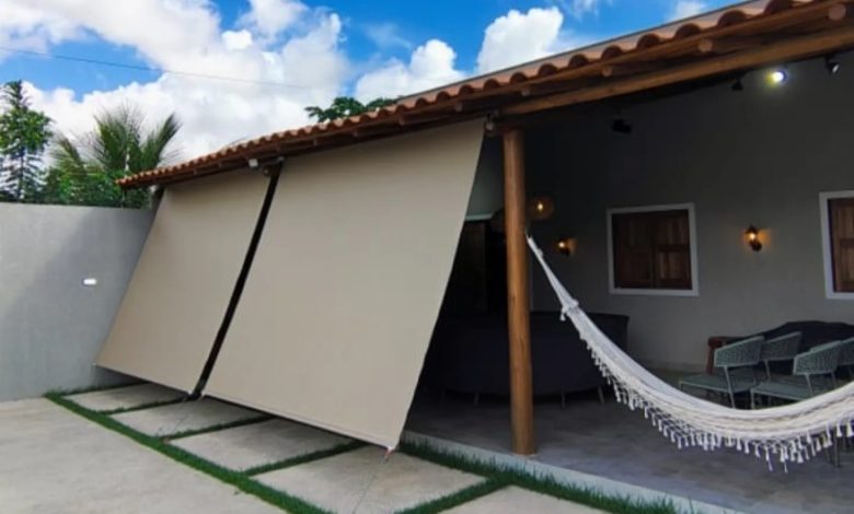 Toldo Cortina retrátil bege sombreando área externa com cadeiras e rede aconchegante.