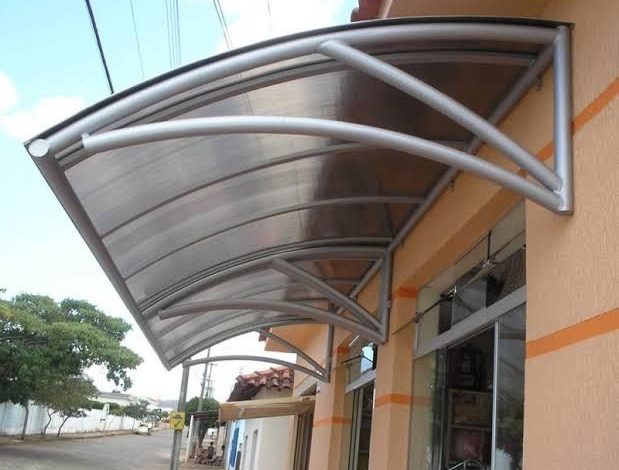 Toldo policarbonato curvo, estrutura metálica moderna instalada sobre entrada comercial.