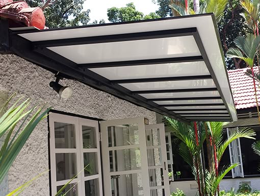 Toldo fixo de policarbonato transparente com estrutura de ferro sobre entrada.