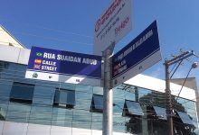 Placa de rua trilíngue com bandeiras e prédio moderno ao fundo.