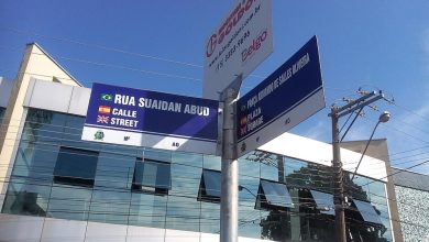 Placa de rua trilíngue com bandeiras e prédio moderno ao fundo.