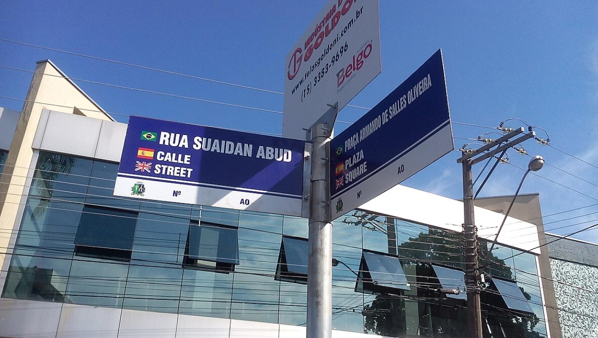 Placa de rua trilíngue com bandeiras e prédio moderno ao fundo.