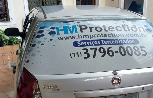 Carro com adesivo perfurado exibe marca HM Protection e contatos.